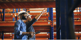 Two workers look at a clipboard and a tablet in front of crates of plants in a warehouse facility