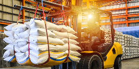 Forklift lifting a large bundle of white sacks secured with straps inside a warehouse with stacked inventory.
