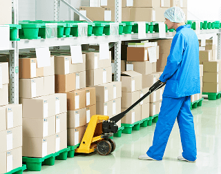 A worker wearing blue safety clothes and a hairnet uses a hand pallet jack to move a pallet of products from a shelving unit