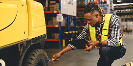 Warehouse worker inspecting a forklift tire and taking notes on a clipboard during a safety check.