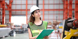Worker wearing a hard hat and safety vest taking notes on a clipboard inside a manufacturing facility.