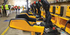 A row of yellow electric forklifts parked and charging as a group of workers walks behind them