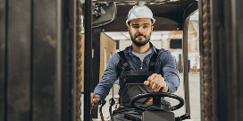 A worker in a hard hat drives a forklift in a warehouse setting