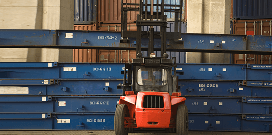 A red forklift picking up a large steel beam from a pile of beams in from of other shipping crates